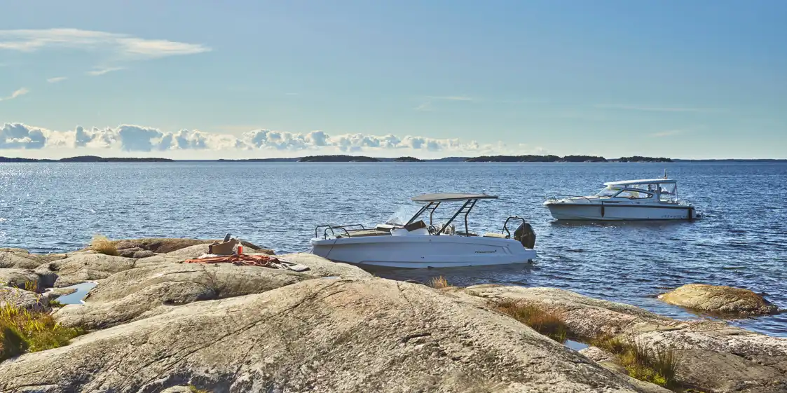 Alla-sorters-batar-1080x540 Två motorbåtar ligger förtöjda vid en solig klippö i skärgården, med hav och öar i bakgrunden.