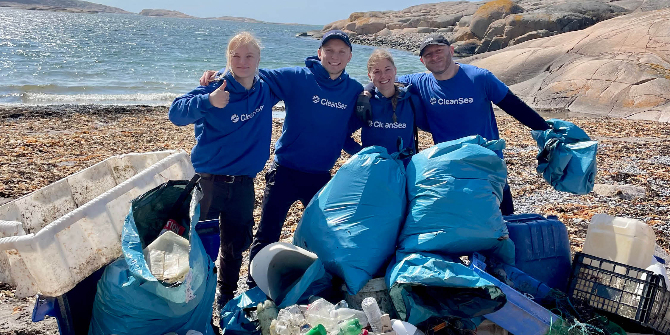 Fyra personer i blå tröjor med texten "CleanSea" poserar på en stenig strand med armarna runt varandra. Framför dem ligger stora blå soppåsar fyllda med uppsamlade skräp, samt plastföremål som flaskor, en korg och en bit isolering. Havet syns i bakgrunden under en solig himmel.