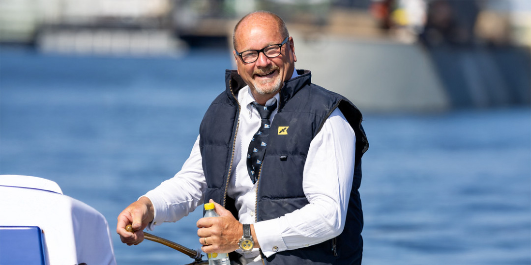 ÖA Stadshusregattan_påbyggd bild_1080x540 En leende man i glasögon och mörk väst över vit skjorta styr en båt på vattnet. Han håller i ratten med ena handen och en flaska med den andra.